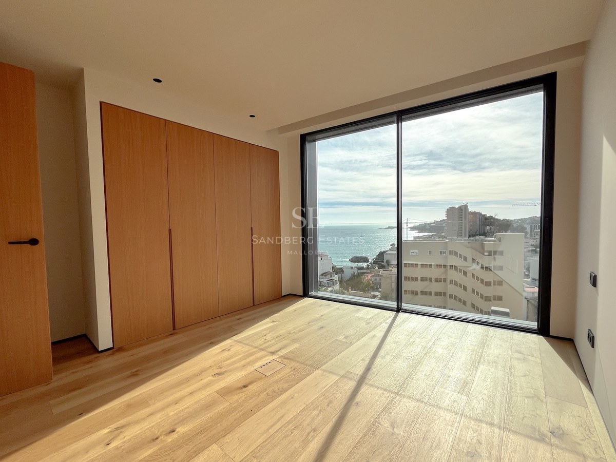 Bright modern bedroom with floor-to-ceiling glass windows overlooking the sea, built-in oak wardrobes, and light wood floors.