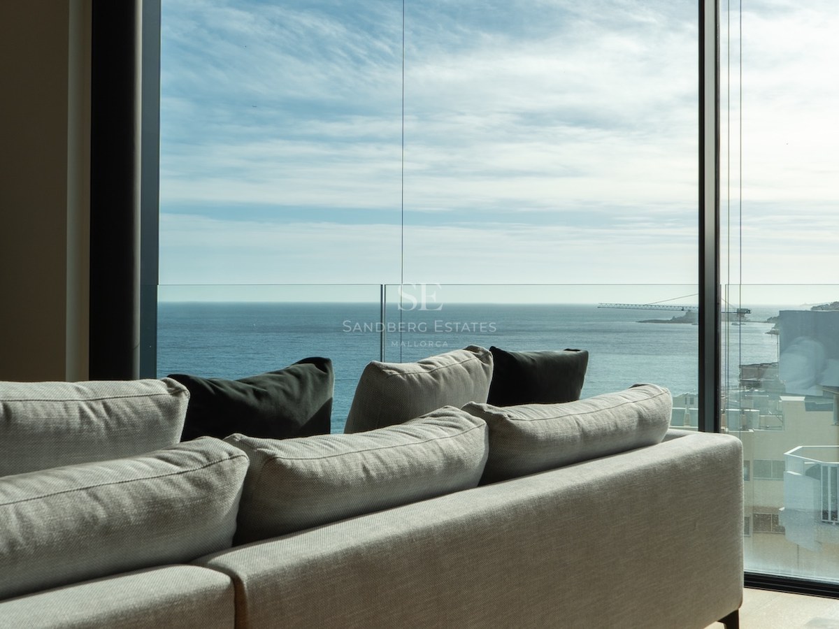 Modernes Sofa vor bodentiefen Glasfronten mit weitem Blick auf das Mittelmeer und blauen Himmel.