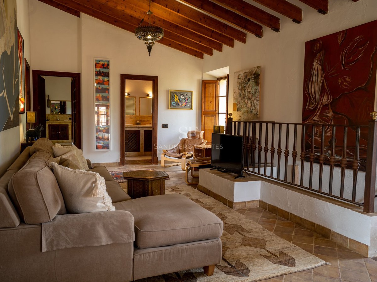 Sala de estar con vigas de madera, suelos de terracota, sofá beige y barandilla de hierro forjado.
