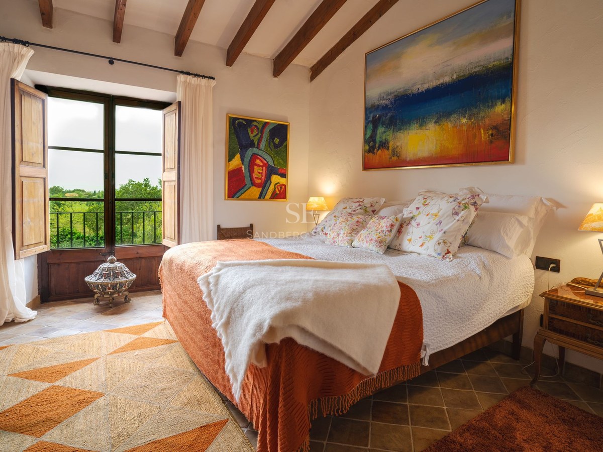 Dormitorio luminoso con vigas de madera expuestas, suelos de terracota y ventanal con vistas a la naturaleza.
