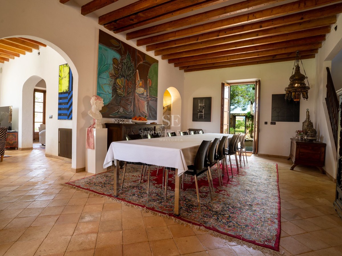 Mesa de comedor amplia sobre alfombra clásica en sala con suelos de terracota y vigas de madera.