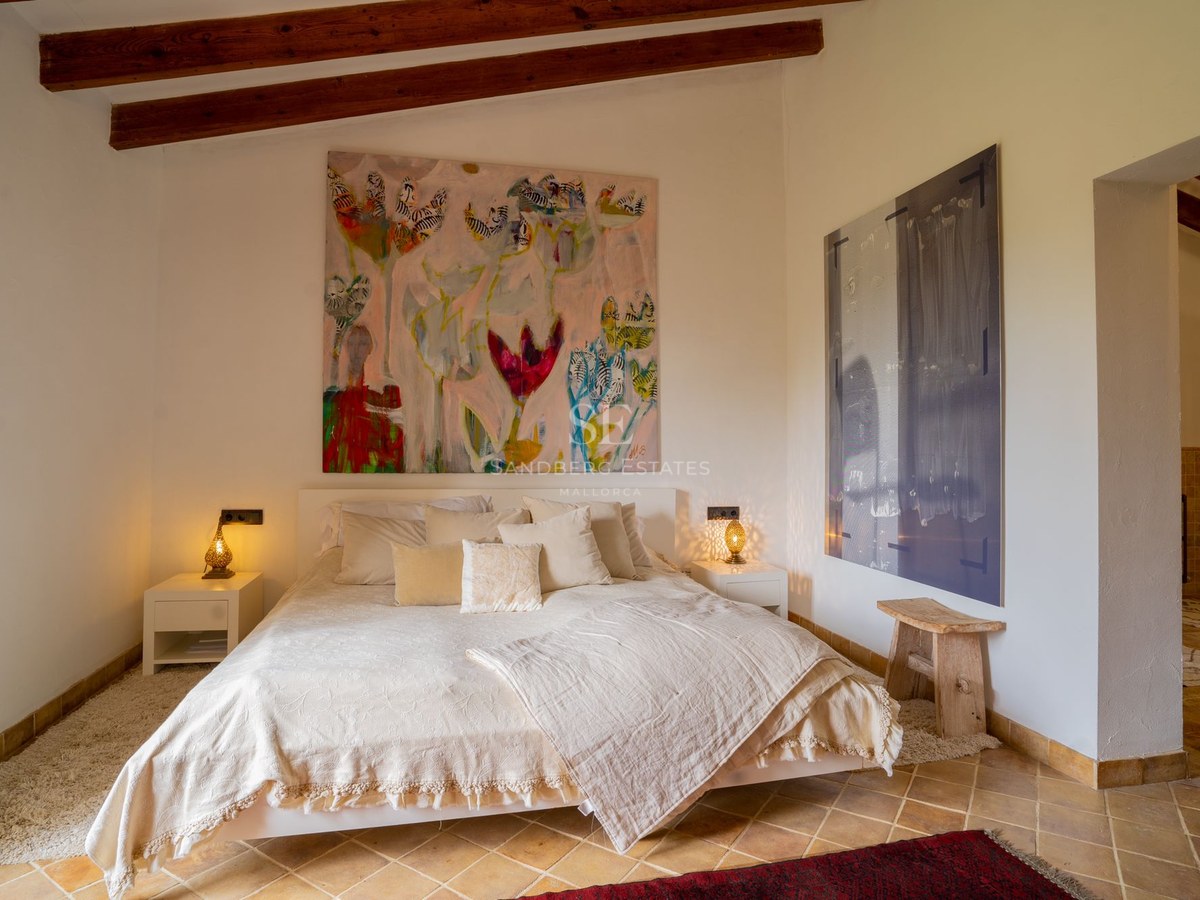 Dormitorio principal con vigas de madera vistas, suelos de terracota, cama amplia y una pintura floral grande.