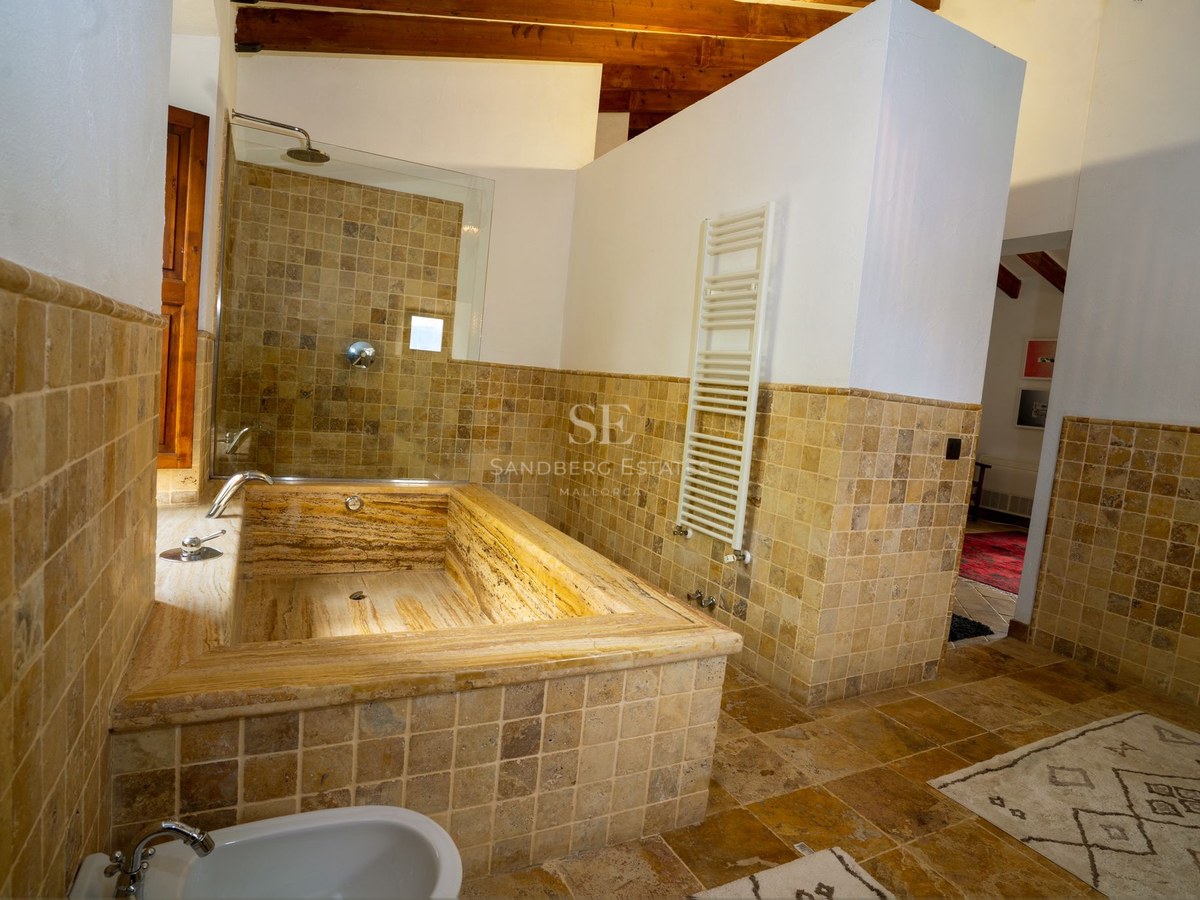 Baño de lujo con bañera integrada de travertino, azulejos de piedra natural y vigas de madera a la vista.