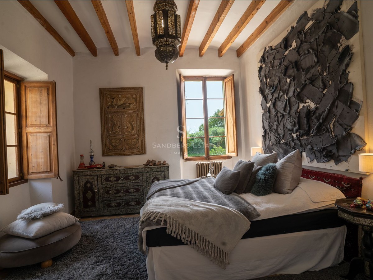 Habitación con vigas de madera vistas, cama amplia con textiles grises y ventana hacia el jardín.