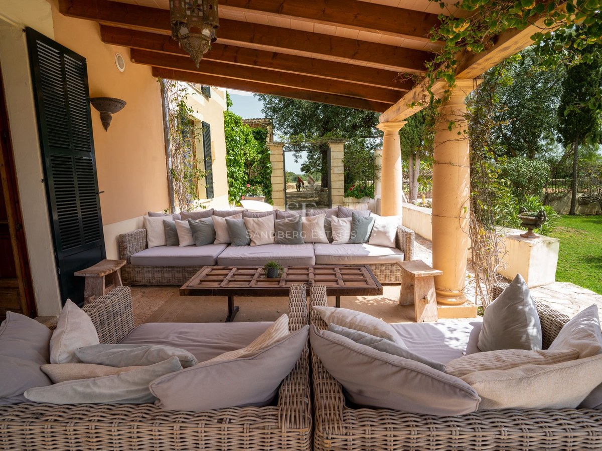Terraza cubierta con sofás de mimbre, vigas de madera vistas y columnas de piedra con vistas al jardín.