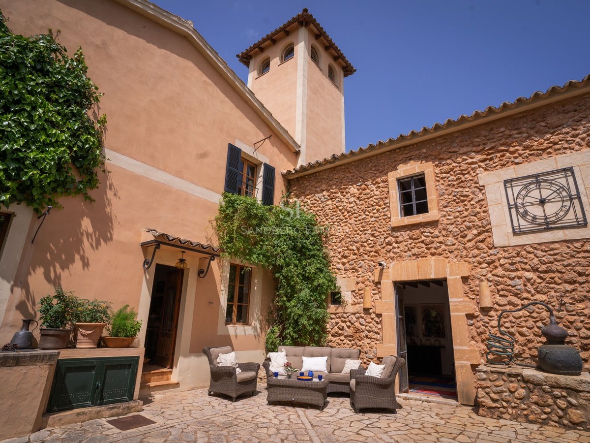 Patio de una villa de piedra con muebles de exterior, paredes de terracota, una torre y hiedra bajo un cielo azul.