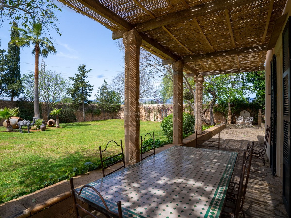 Terraza a la sombra con mesa de mosaico y sillas de hierro frente a un jardín privado con césped y árboles.