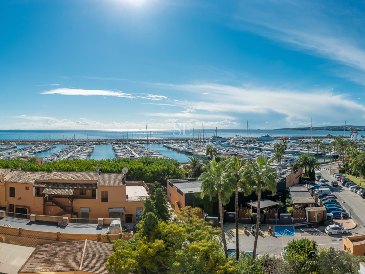 Elevated view of a Mediterranean port with yachts, terracotta buildings, and palm trees under a bright blue sky.