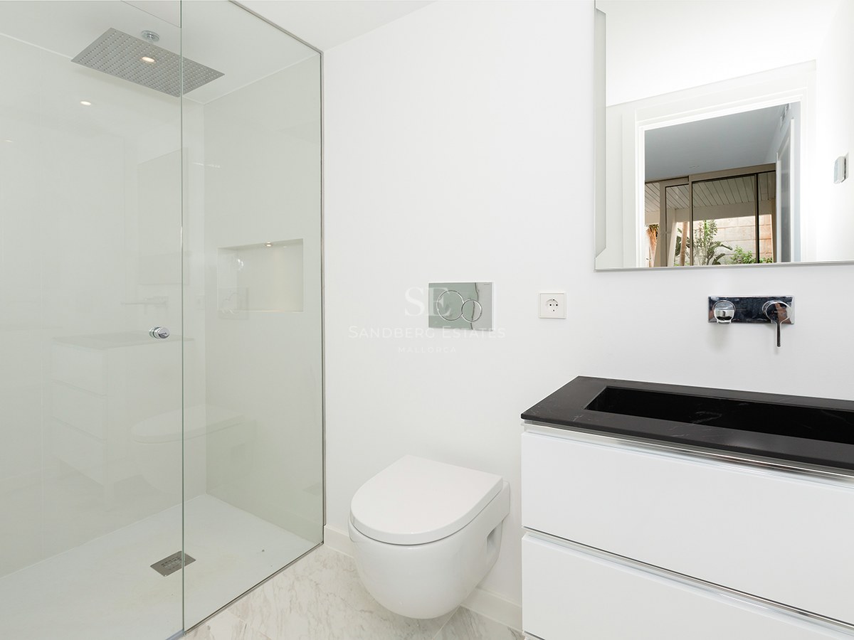 A bright, minimalist bathroom featuring a glass walk-in shower, a white wall-hung toilet, and a black and white vanity.