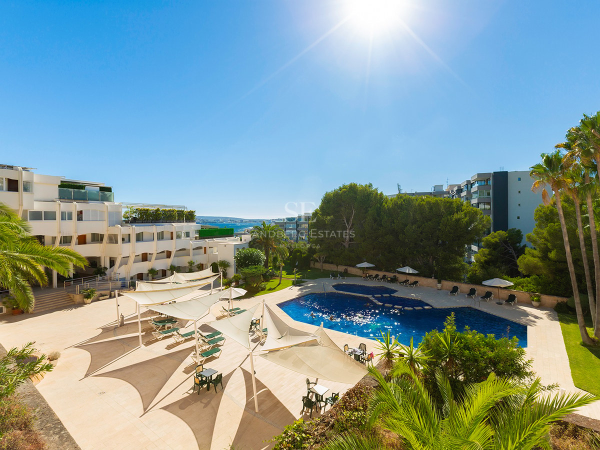 Large blue swimming pool surrounded by beige terrace, white sun sails, palm trees, and resort buildings under a bright sun.