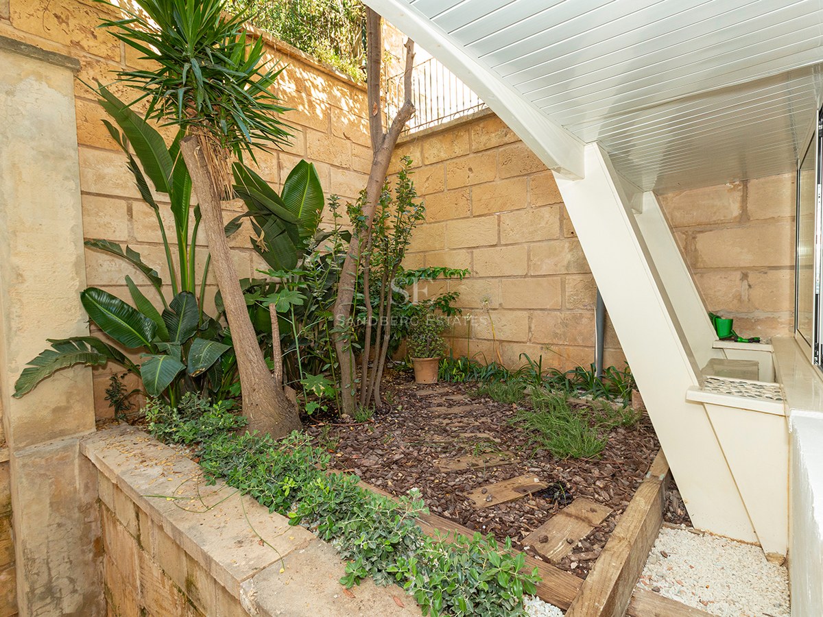 A small outdoor garden area with tropical plants, wood mulch, and a sandstone wall under a slanted white structure.