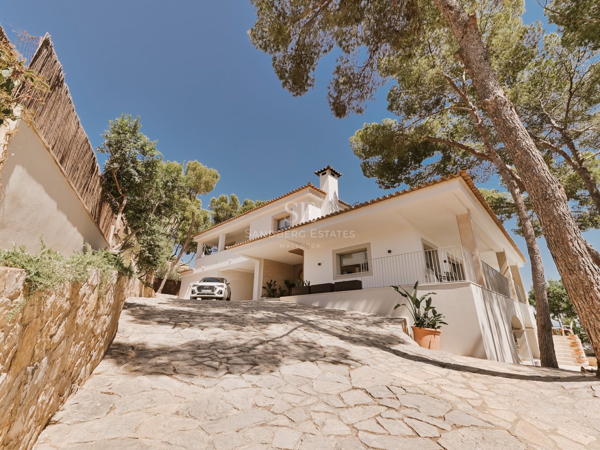 Vista exterior de una villa blanca de estilo mediterráneo con camino de piedra natural y pinos maduros.