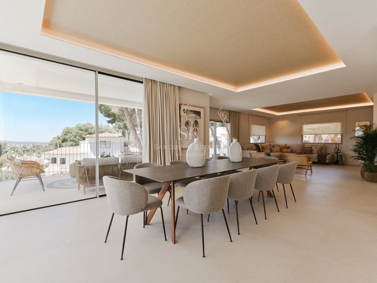 Comedor minimalista con mesa de madera, diez sillas de terciopelo gris y grandes ventanales correderos de cristal.