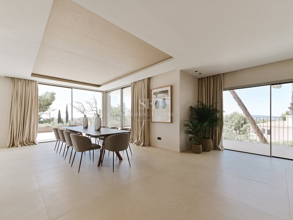 Comedor amplio con mesa para ocho, ventanales de suelo a techo, suelos de piedra clara y luz natural abundante.