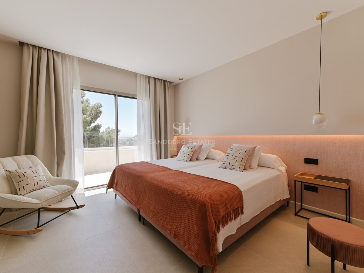 Dormitorio moderno con cama doble, manta terracota y ventanales de suelo a techo que dan a una terraza soleada.
