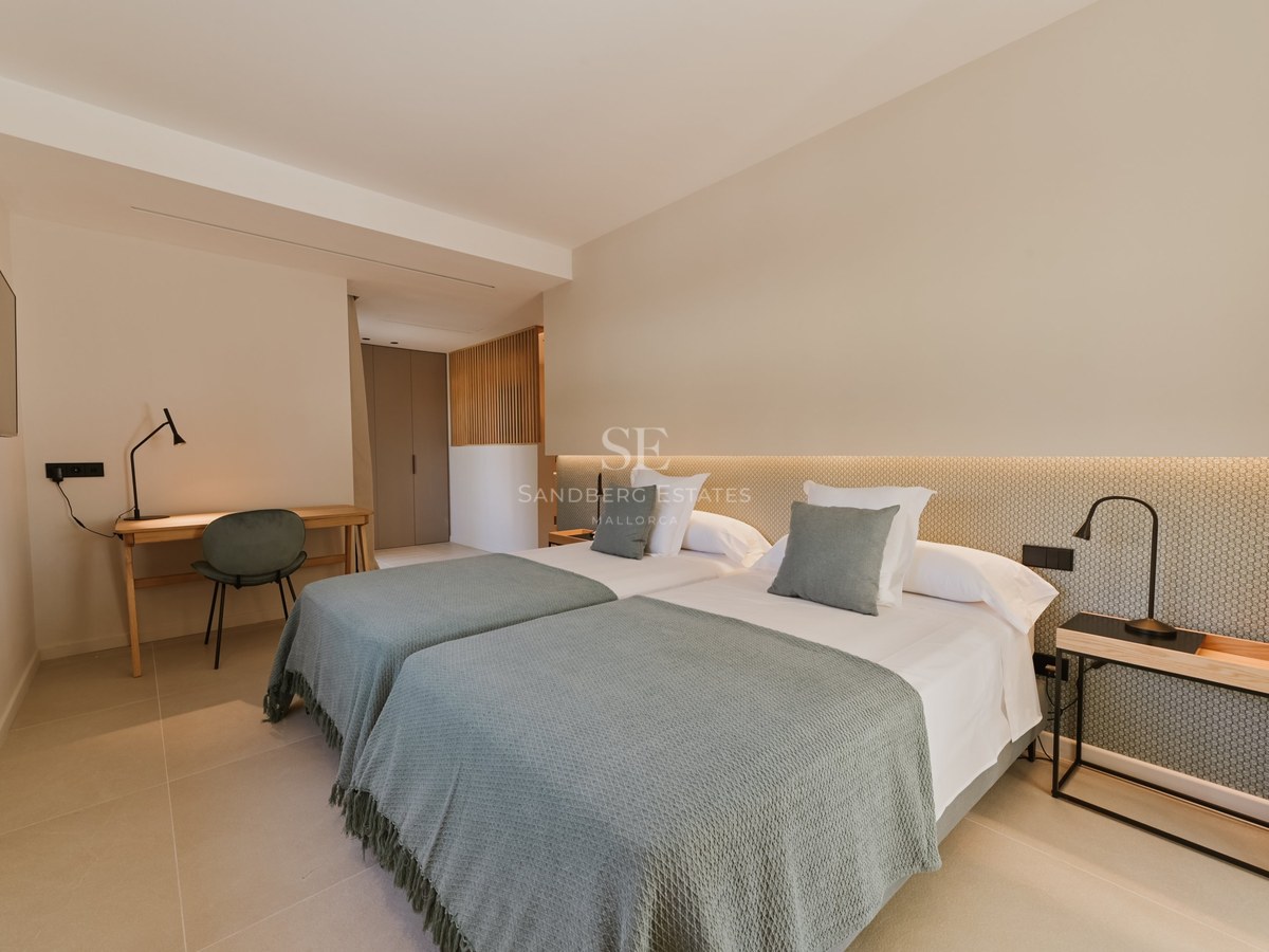 Dormitorio moderno con dos camas individuales, mantas verde salvia, escritorio de madera y luz cálida indirecta.