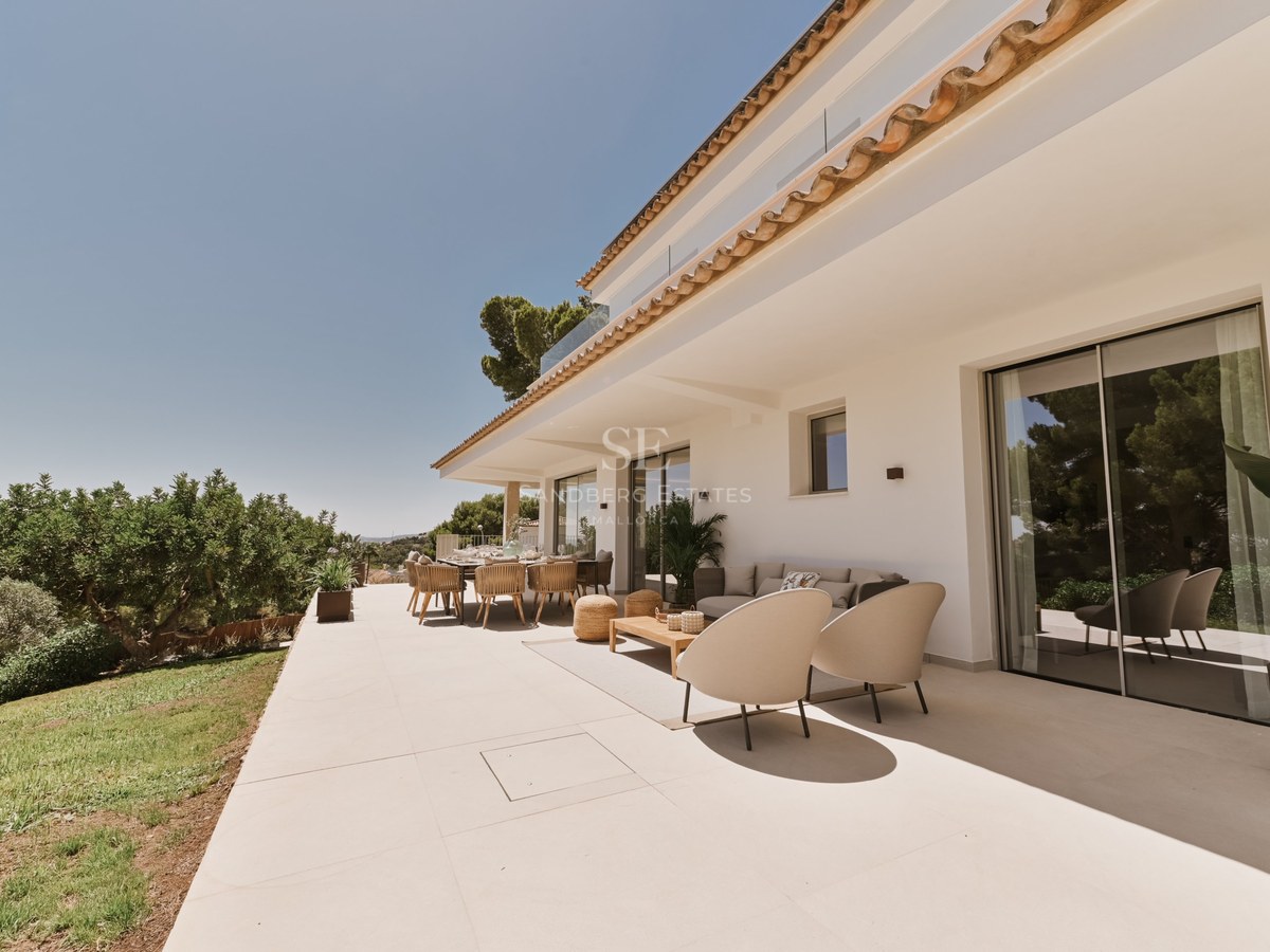 Amplia terraza soleada de piedra con muebles de salón de diseño, zona de comedor y fachada de villa blanca.