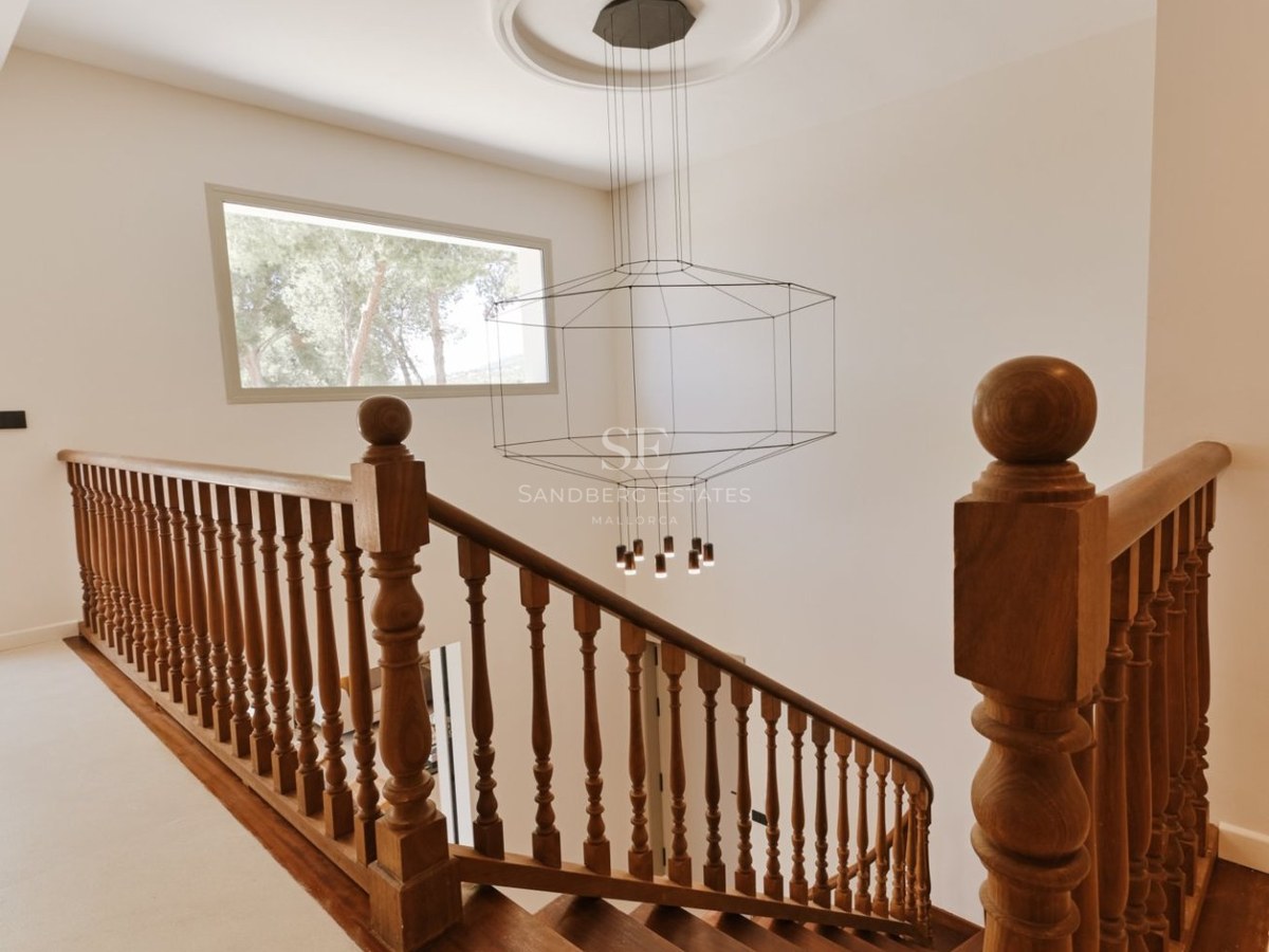 Escalera de madera oscura con barandilla clásica y moderna lámpara de araña de alambre en un pasillo luminoso.