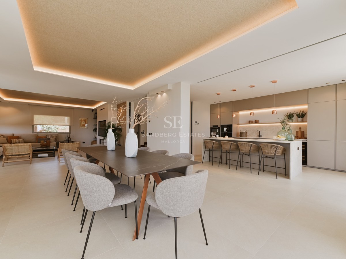Comedor moderno con mesa larga, sillas tapizadas grises y cocina minimalista con iluminación LED indirecta.