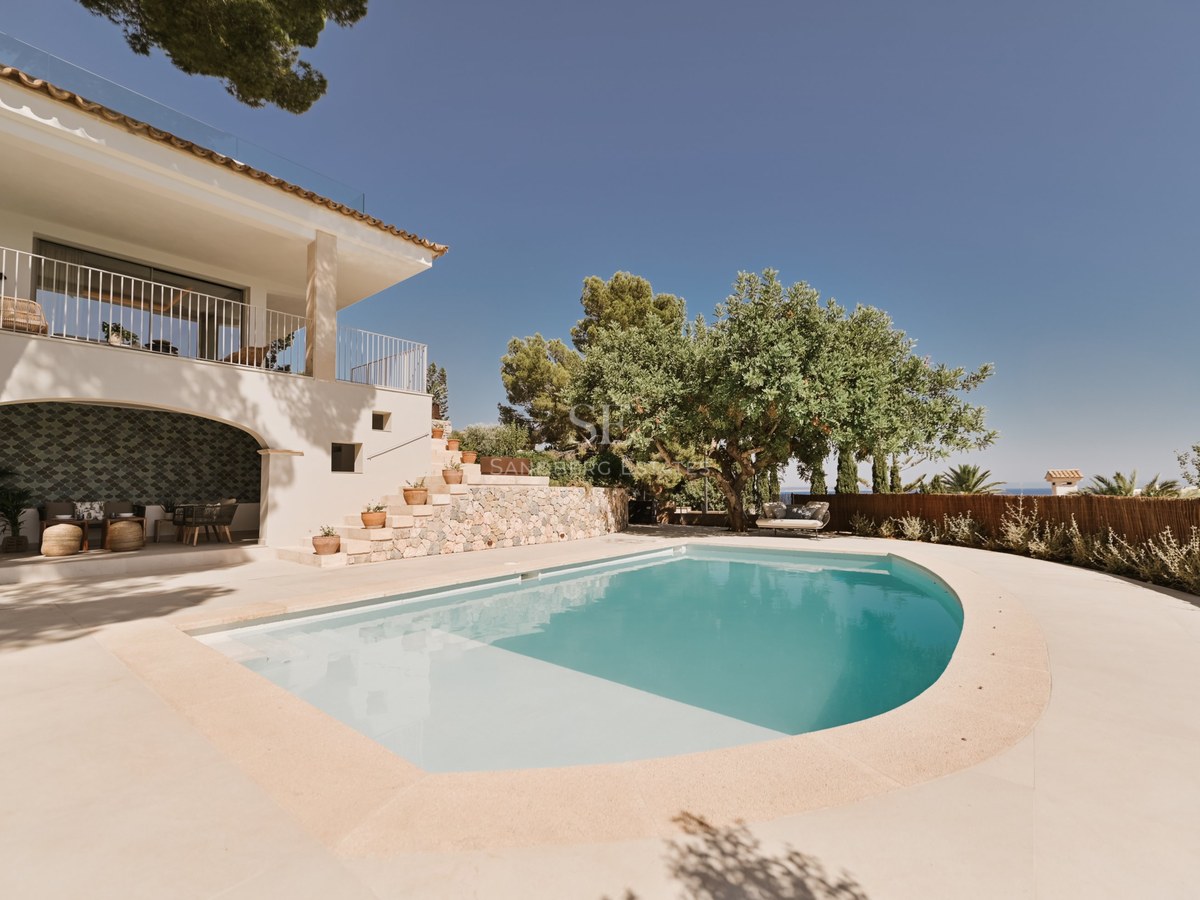Piscina curva turquesa junto a una villa de paredes blancas con escaleras de piedra y vegetación mediterránea.