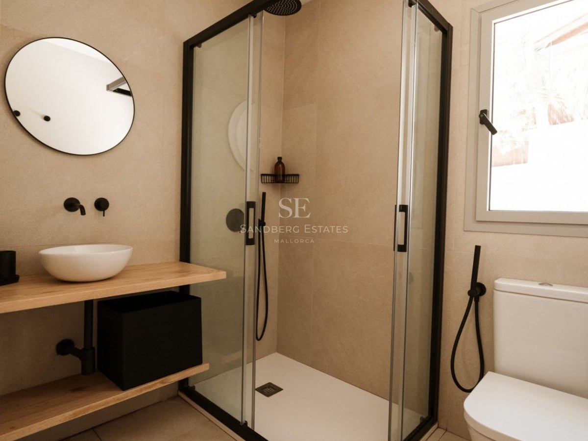 Baño moderno con ducha de cristal, grifería negra mate, encimera de madera y espejo redondo.