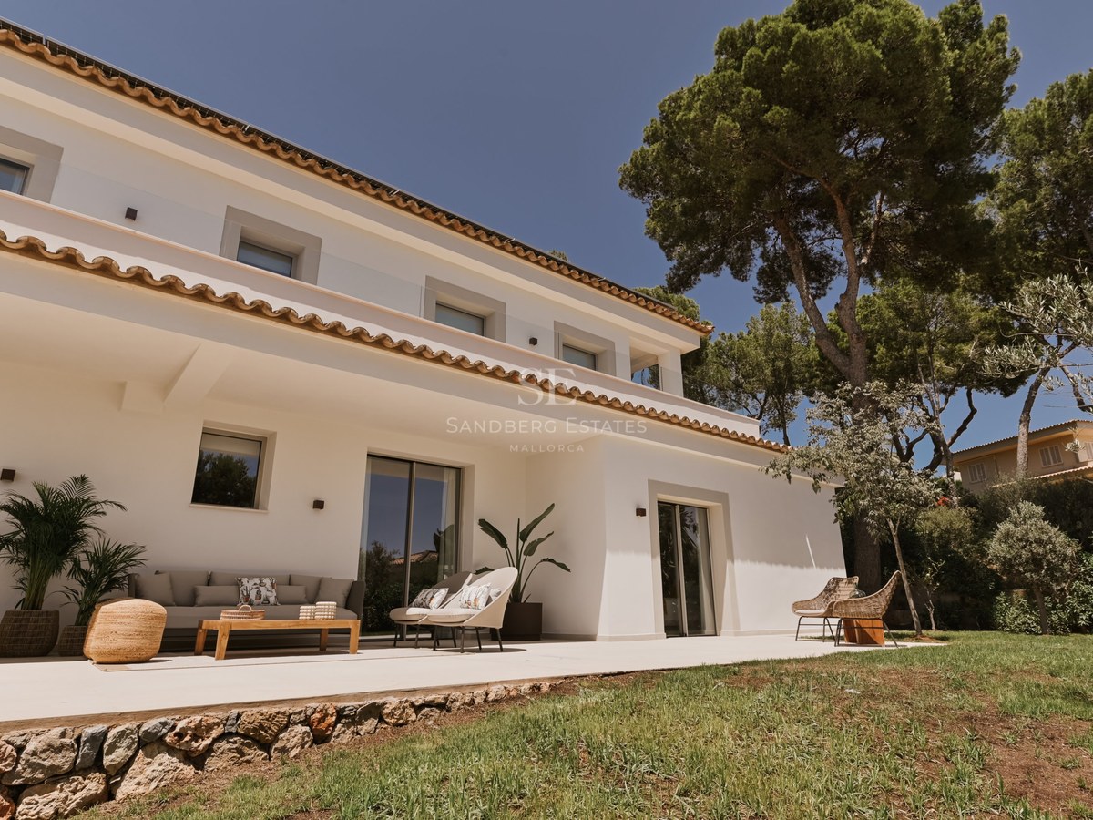 Villa blanca de dos plantas con tejado de terracota, muebles de exterior y pinos bajo un cielo despejado.