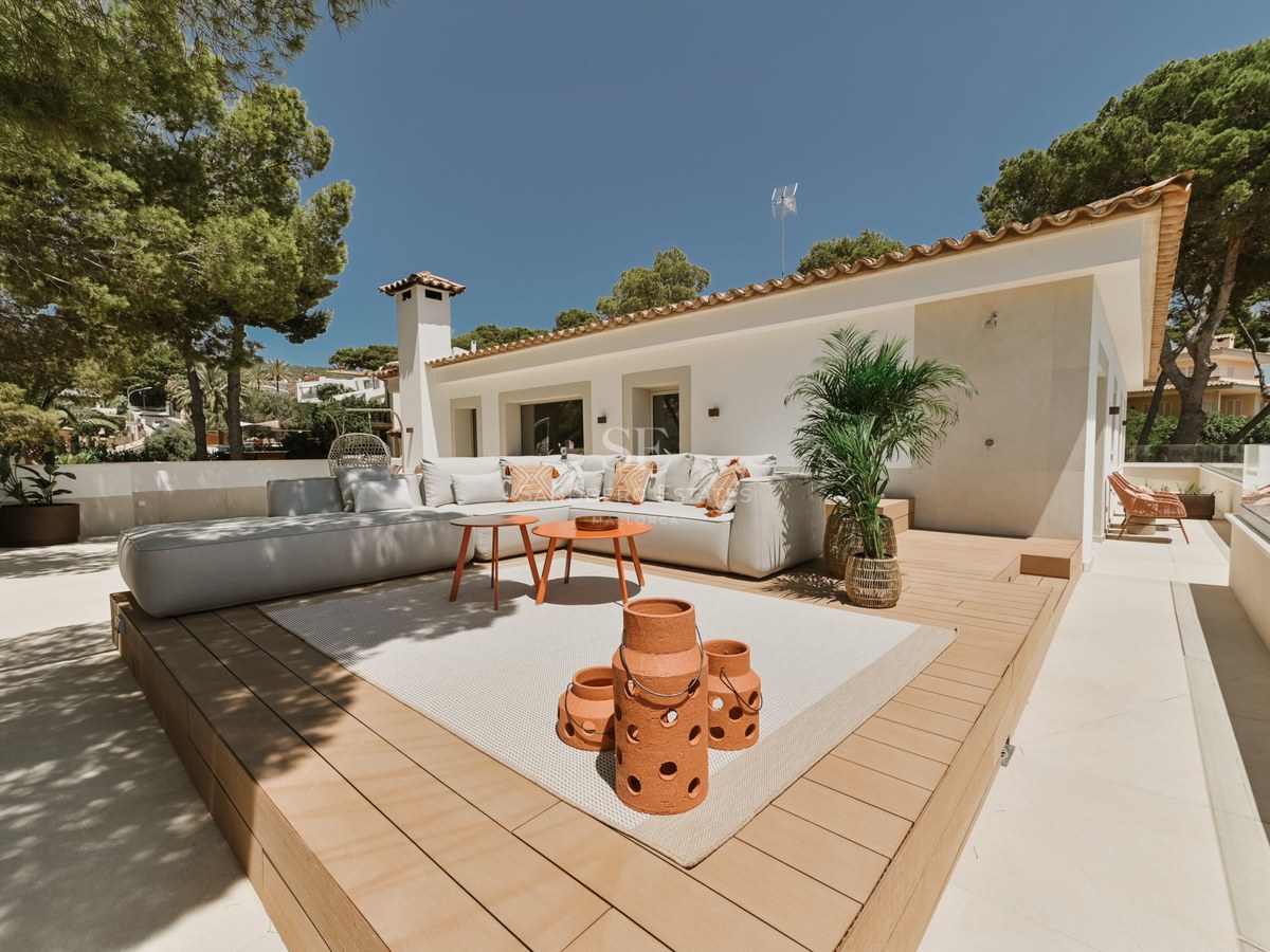 Terraza soleada con sofá en L blanco sobre tarima de madera, linternas de terracota y fachada de villa blanca.
