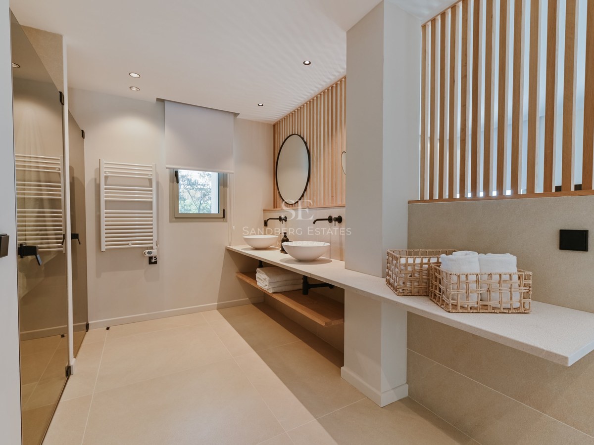 Baño moderno con tocador doble, lavabos sobre encimera, pared de listones de madera y espejo redondo.