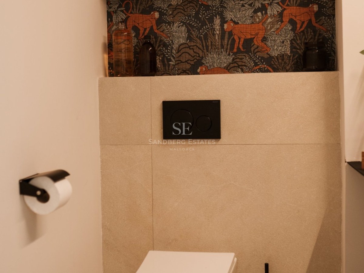 Inodoro suspendido moderno con pulsador negro, azulejos beige y hornacina decorativa con papel pintado de monos.