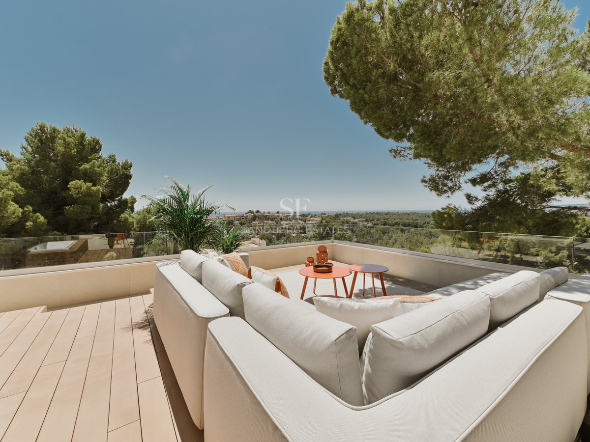 Sofá modular blanco en terraza de madera con barandillas de cristal y vistas al paisaje mediterráneo y al mar.