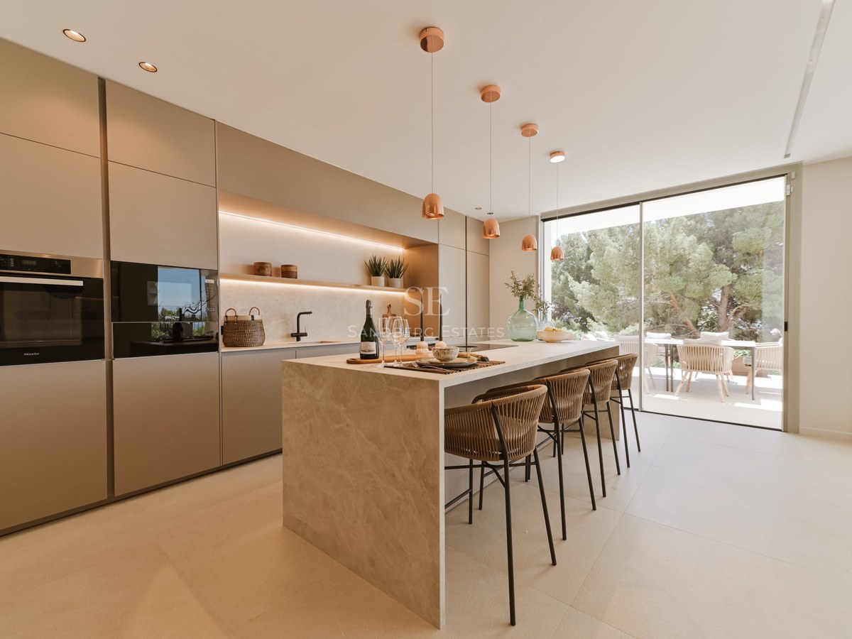Cocina minimalista moderna con isla de piedra beige, taburetes y ventanales de suelo a techo con vistas al jardín.