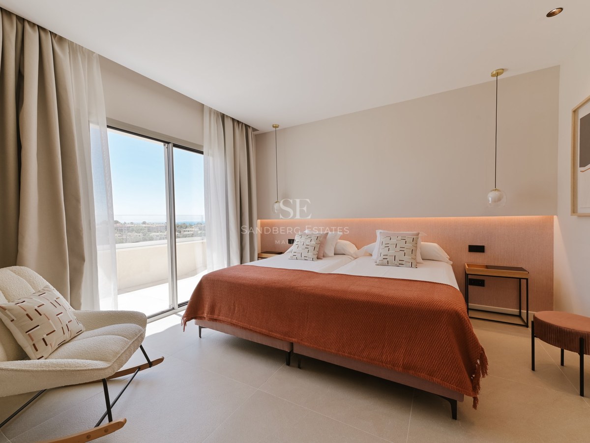 Dormitorio moderno con cama terracota, iluminación indirecta y ventanales de suelo a techo hacia una terraza soleada.