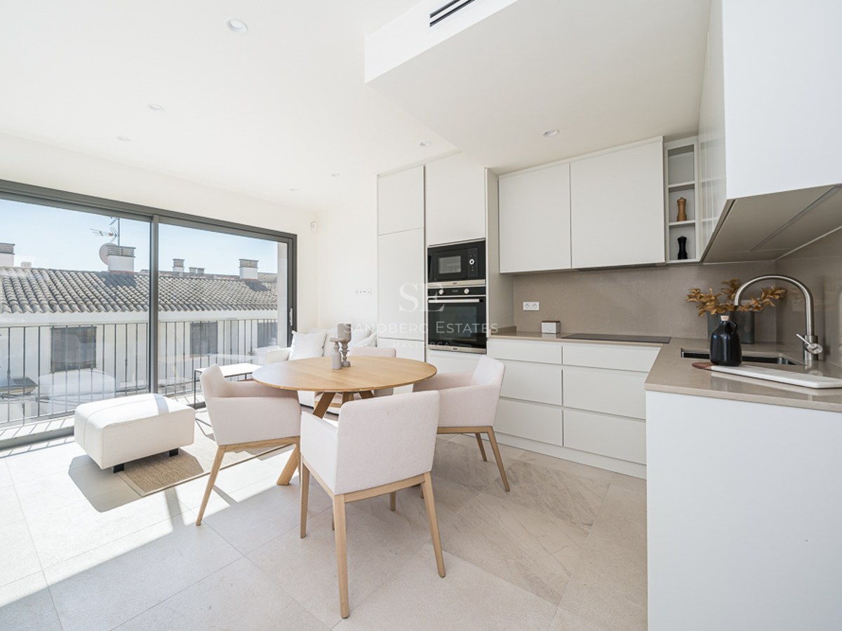 Cocina luminosa con muebles blancos, mesa de comedor de madera y grandes ventanales con vistas a tejados tradicionales.