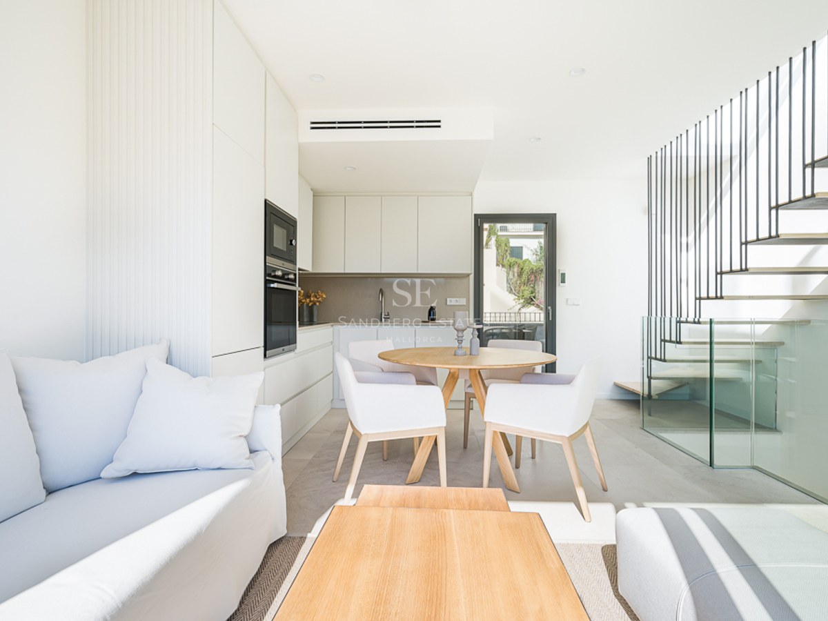 Salón minimalista luminoso con sofá blanco, mesa de comedor de roble, cocina moderna y escalera flotante con cristal.