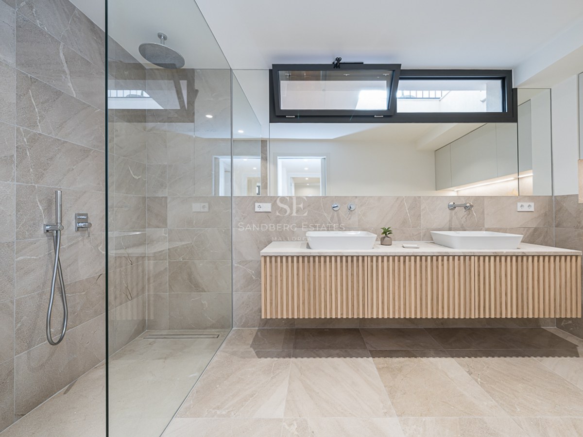 Baño moderno con ducha de cristal, azulejos de piedra color arena y mueble doble suspendido de madera.