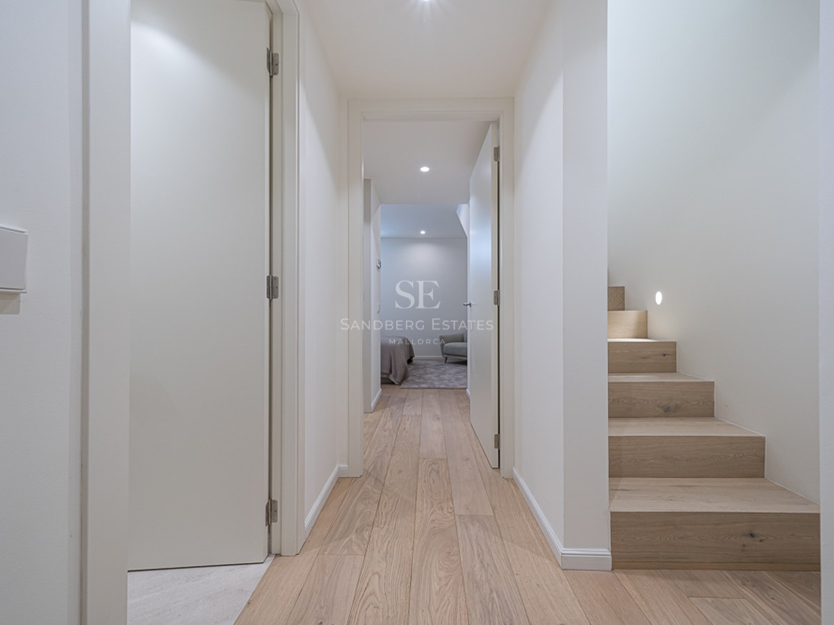 Pasillo moderno con suelos de madera clara, paredes blancas y escalera de madera con iluminación integrada.