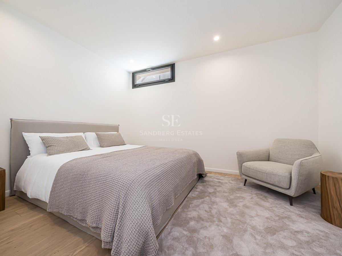 Dormitorio moderno con cama tapizada, mesitas de madera natural, sillón gris y suelos de roble claro.