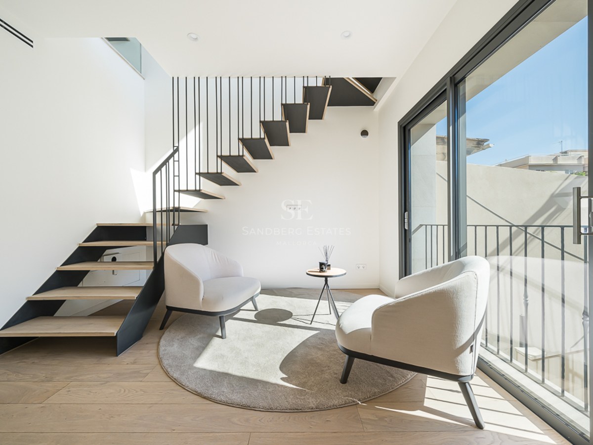 Espacio moderno con escalera flotante de madera, dos sillones crema y ventanales de suelo a techo.