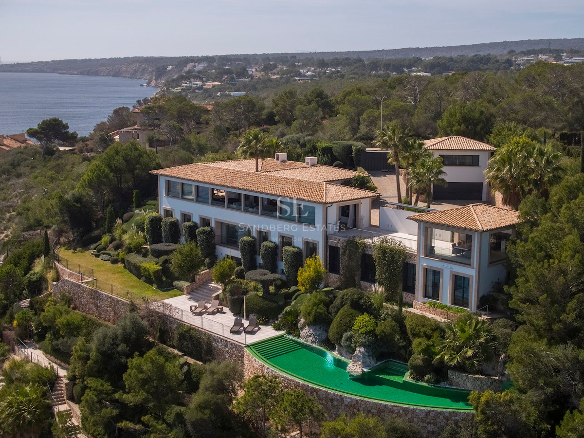 Aerial view of a white multi-level luxury villa with a green infinity pool overlooking the Mediterranean coast.