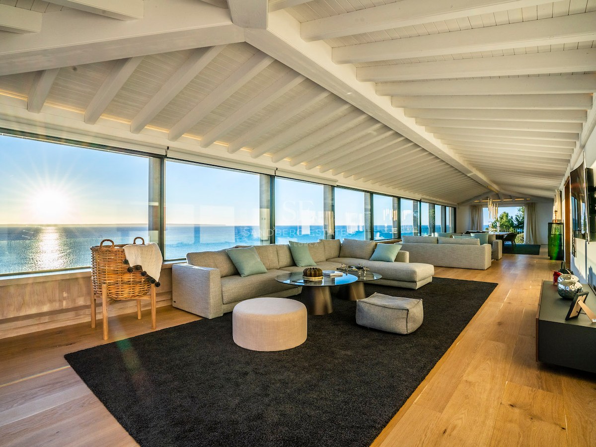 Modern living room with white beamed ceilings, oak floors, and floor-to-ceiling windows overlooking the ocean at sunset.
