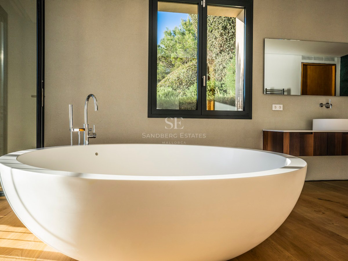 Modern master bathroom featuring a large white oval freestanding tub on oak floors with garden views through a window.