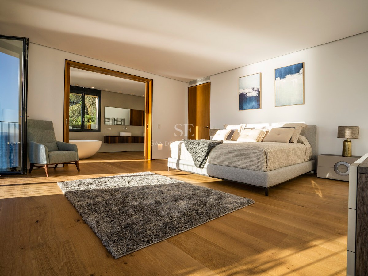 Modern master bedroom with oak floors, king-size bed, sea-view terrace access, and open-plan en-suite bathroom.