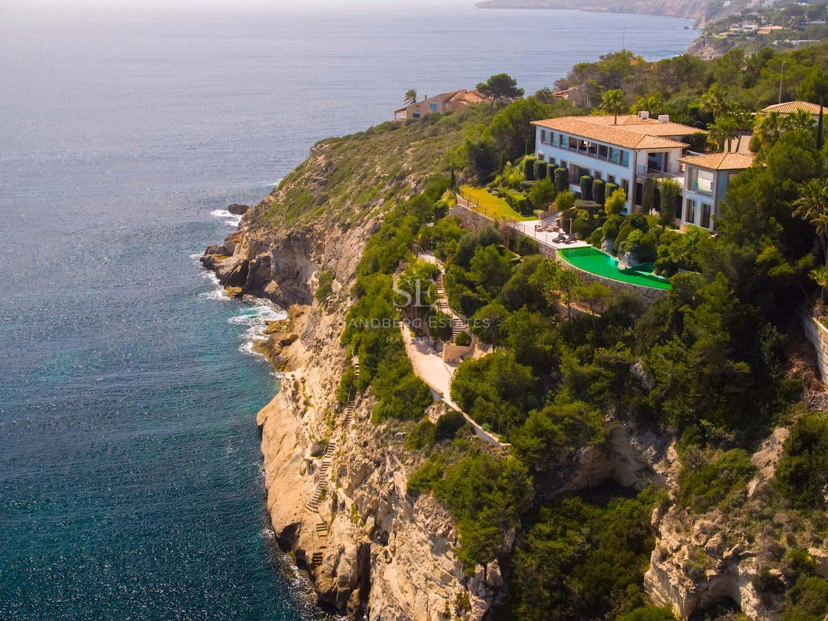 Aerial view of a luxury villa perched on a Mediterranean cliff with an emerald pool and expansive ocean views.