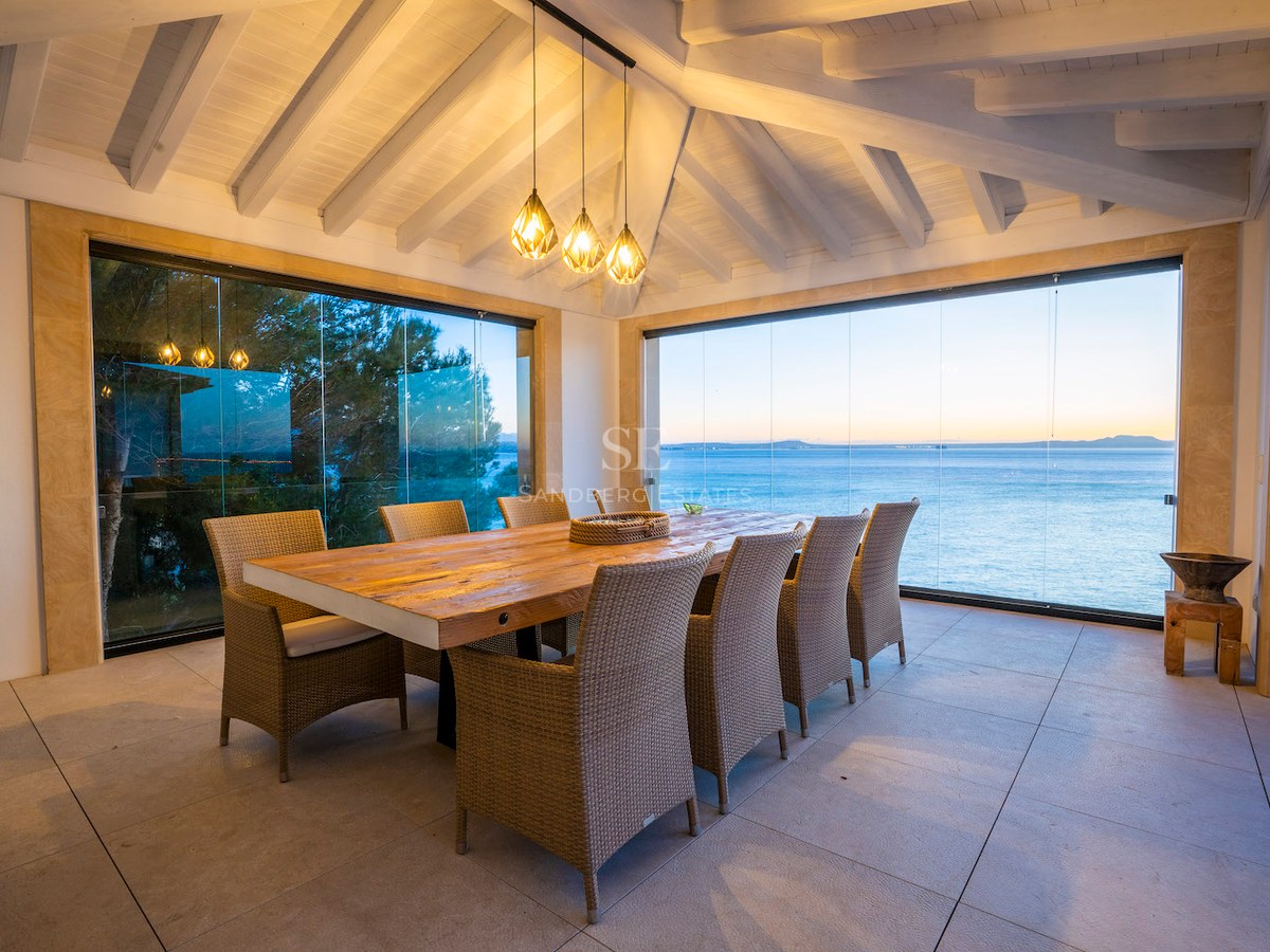 Large wooden dining table on a terrace with glass walls overlooking the blue sea at dusk under a beamed ceiling.