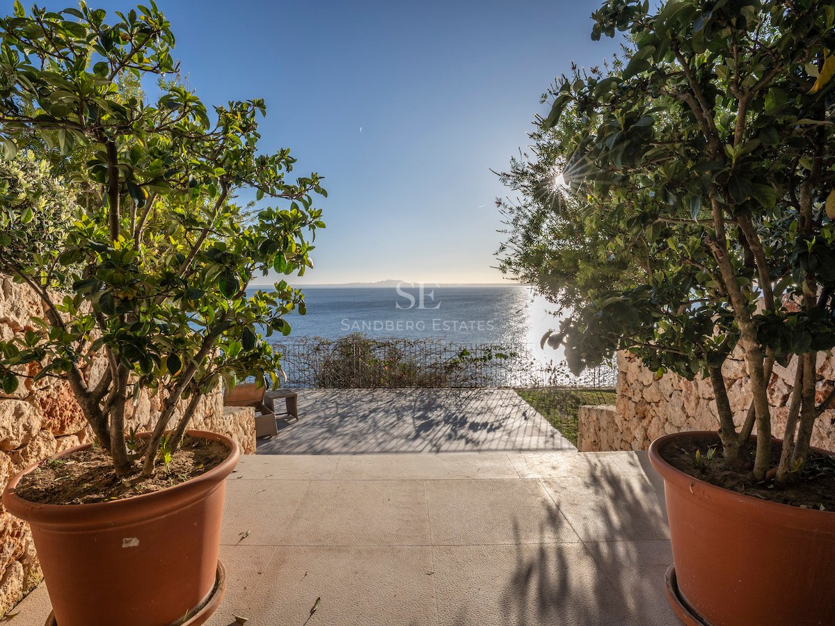 View of the blue sea from a stone terrace framed by potted plants and dry stone walls under a clear sky.