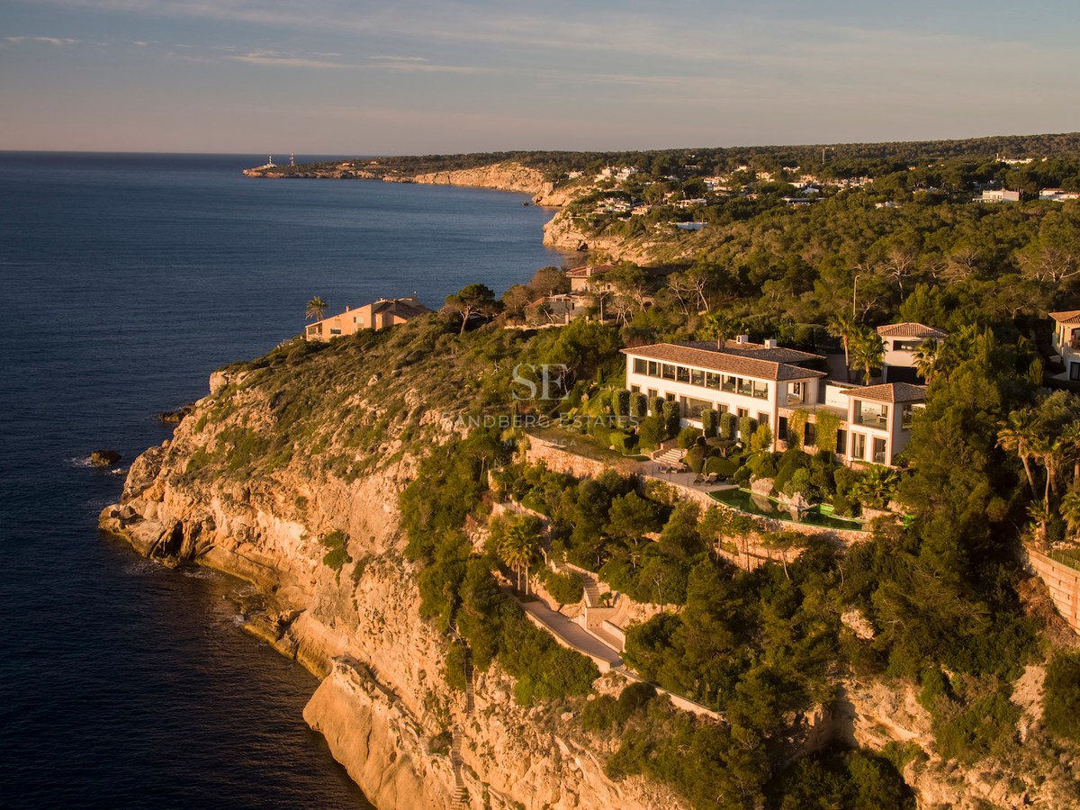 Aerial drone shot of a luxury white villa on a rocky cliff edge overlooking the Mediterranean sea at golden hour.