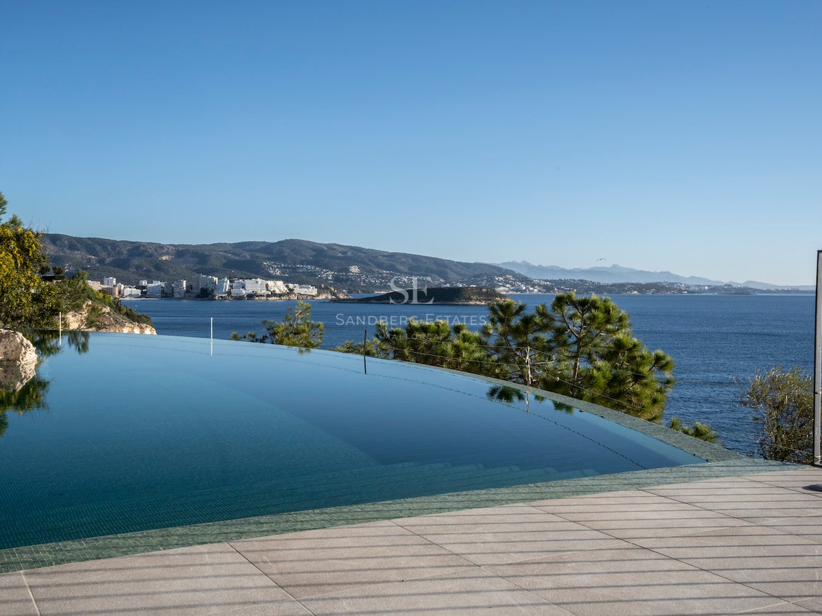 An infinity swimming pool with clear blue water overlooking the sea and a mountainous coastline under a clear sky.
