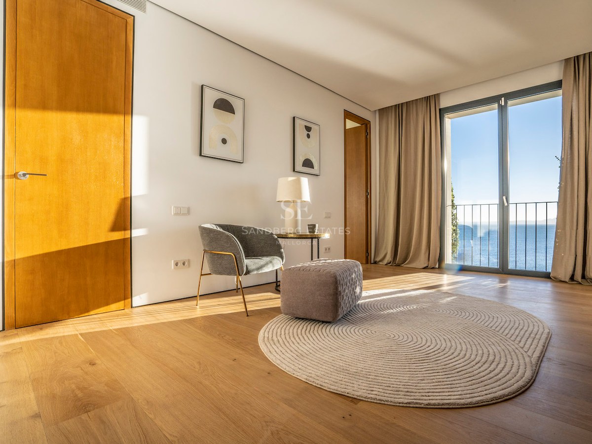Modern room with oak floors, grey armchair, and glass doors opening to a Mediterranean sea view.