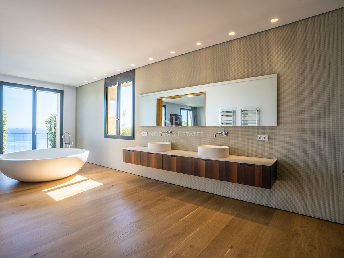 Modern bathroom with freestanding oval bathtub, oak floors, double vanity, and sea views through a glass door.