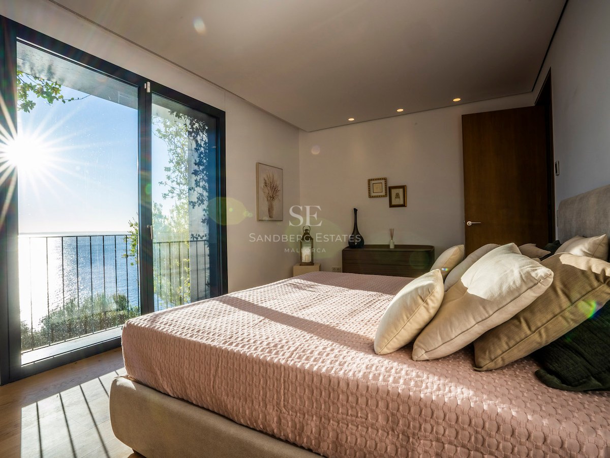 Bright bedroom with a large bed, pink linens, oak floors, and a floor-to-ceiling window overlooking the blue sea.
